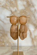 A pair of chunky statement earrings with large, rounded rectangular amber-orange stone tops and cascading layered petal-shaped drops in a matching warm tan hue, set in polished gold-tone metal and displayed on a gold stand.