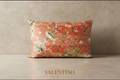 A square decorative pillow featuring a vibrant orange-red background with an intricate vintage-style botanical and scroll pattern in green, beige, and white, displayed on a wooden surface.